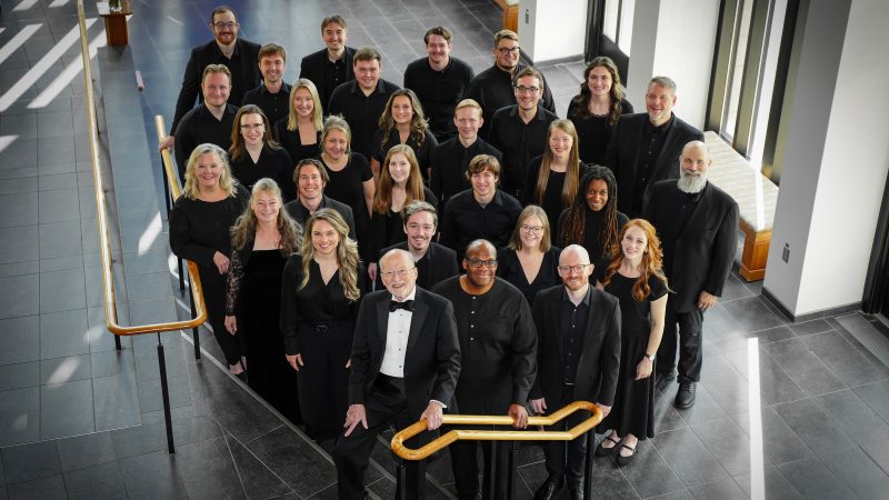 VocalEssence Ensemble Singers wearing black pose for a photo in the lobby of a building with windows and stairs with gold railing. Photo Credit: Novart Media