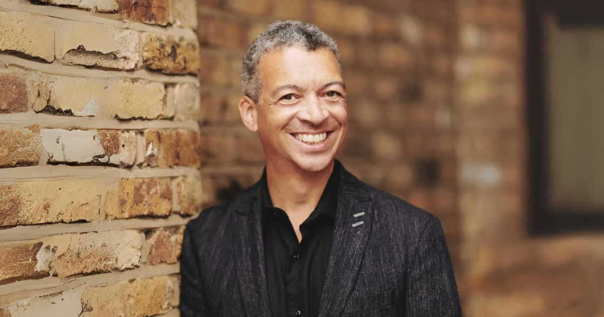 Roderick Williams, wearing a black button down shirt and a dark blazer with a full smile stands in front of a brick wall. Photo Credit: Theo Williams