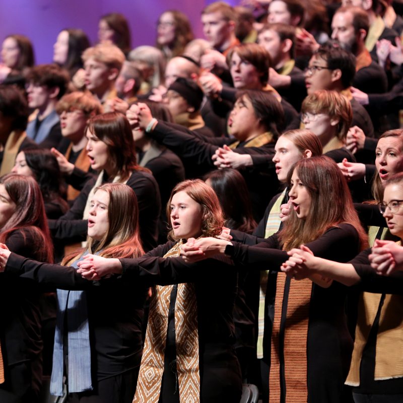 VocalEssence singers wearing black with multicolored stoles perform while holding their arms forward on stage for the annual WITNESS concert. Photo Credit: Kyndell Harkness