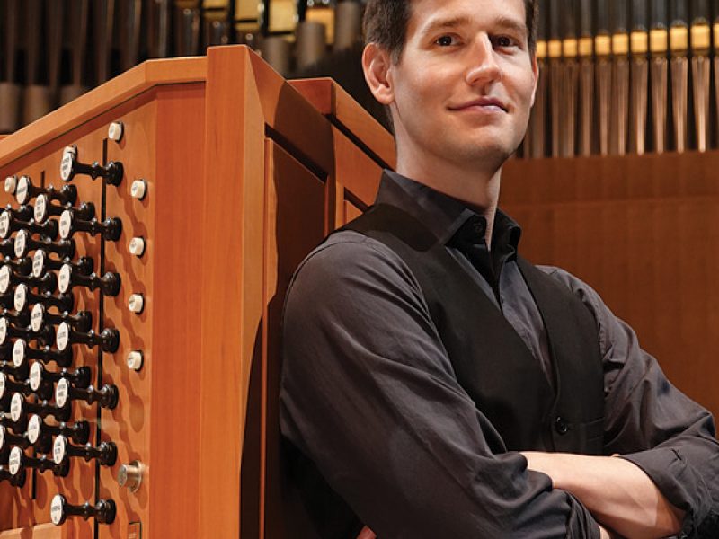 Greg Zelek, wearing a black shirt and vest with hands folded poses in front of an organ console and pipes. Photo Credit: Peter Rodgers