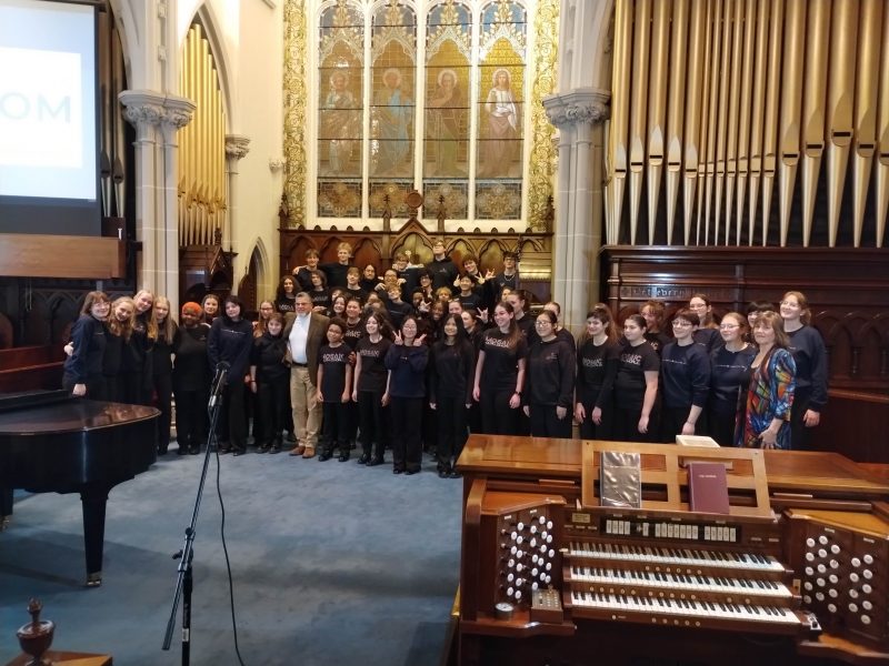 Group of singers standing in the sanctuary of a church in front of organ pipes and a stained glass window.