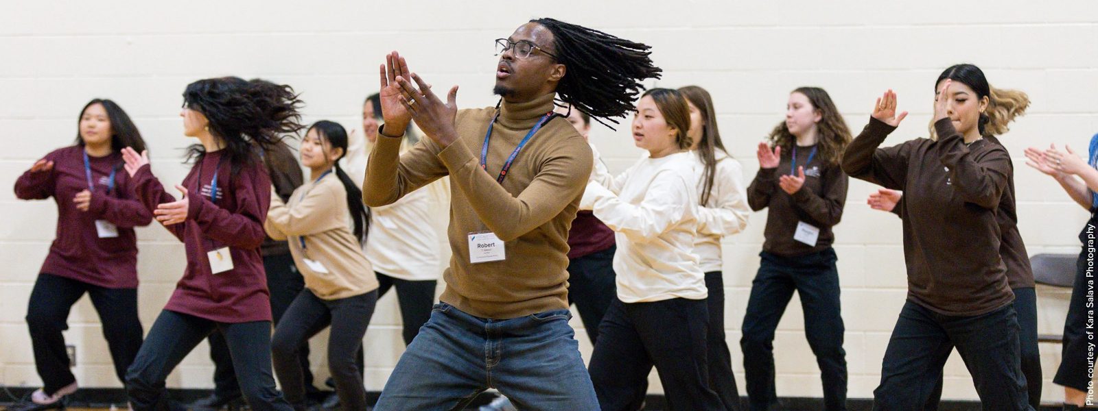 Teenage participants in the Urban Choir Festival wearing long sleeved shirts of different colors and black pants while clapping and dancing led by an instructor with facial hair and glasses wearing a tan turtleneck and jeans clapping and dancing. Photo Credit: Kara Salava Photography.