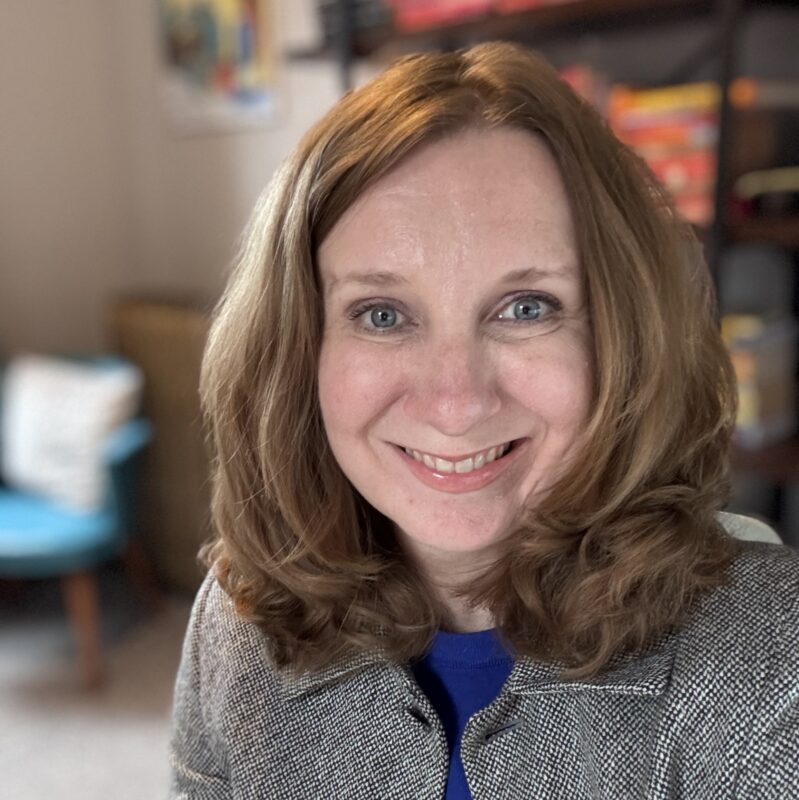 Image of person smiling with shoulder length brown hair wearing a bright blue shirt under a black and white jacket. Photo credit: Jennifer Dieter