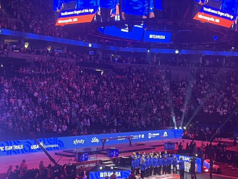 Members of VocalEssence Singers Of This Age, wearing blue shirts and black pants, perform at the U.S. Olympic Trials on a stage and projected on a jumbotron.